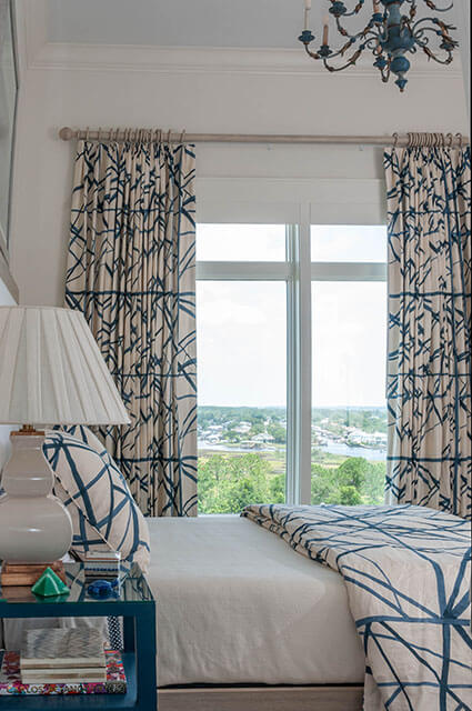 A bright bedroom featuring a large window with a scenic marsh view. The bed has white linens and a blue-and-cream geometric patterned duvet. Matching patterned curtains hang from a light wood rod. A white gourd-style lamp sits on a teal nightstand in the foreground.