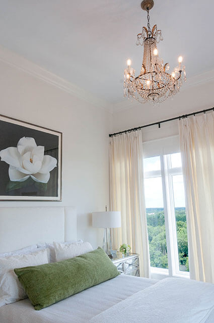 An elegant white bedroom with high ceilings and a crystal chandelier. A large framed print of a white magnolia hangs above a white tufted headboard. A long green lumbar pillow accents the white bedding, and cream-colored curtains frame a view of lush green treetops.