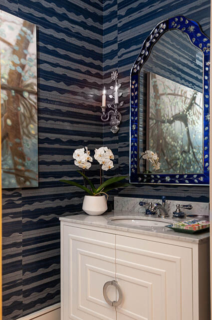 A luxurious powder room with deep blue and grey horizontal-striped wallpaper. It features a white vanity with a marble countertop and a unique arched mirror with a decorative blue and silver floral frame. A white orchid in a ceramic pot sits next to the sink.