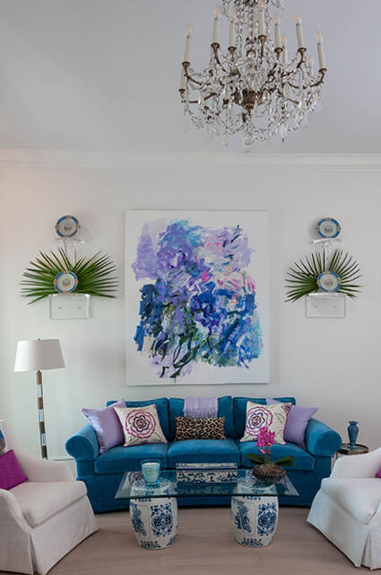 A vibrant sitting area featuring a bright teal sofa adorned with purple and floral pillows. Above the sofa hangs a large abstract painting in shades of blue and lavender. The room includes a glass-top coffee table supported by two blue-and-white porcelain garden stools and an ornate crystal chandelier.