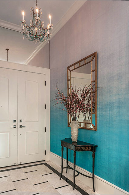 An entryway featuring a dramatic teal-to-lavender ombre textured wallpaper. A slim black console table with a Greek key pattern sits under a gold-framed mirror. A tall glass vase with flowering pink branches provides a focal point next to white double doors and a marble tile floor.