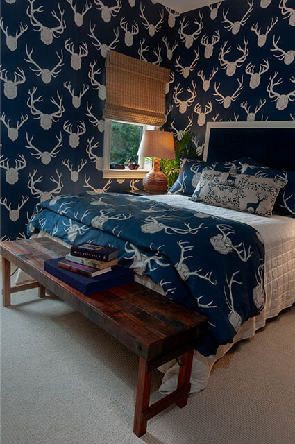 A cozy bedroom with bold navy blue wallpaper featuring a white deer antler pattern. The bed is dressed in a matching navy antler-print duvet and a rustic wooden bench sits at the foot. A warm lamp on the nightstand creates a soft glow against the dark walls.