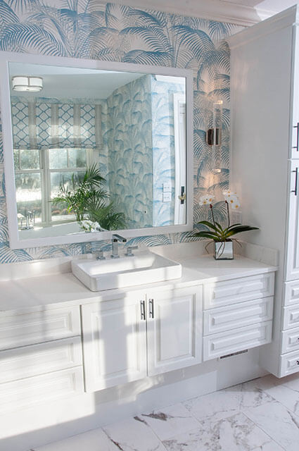 A bright bathroom vanity area featuring light blue palm leaf wallpaper. A large white-framed mirror hangs over a modern white vanity with a rectangular vessel sink. The room is filled with natural light, highlighting the marble tile floors.