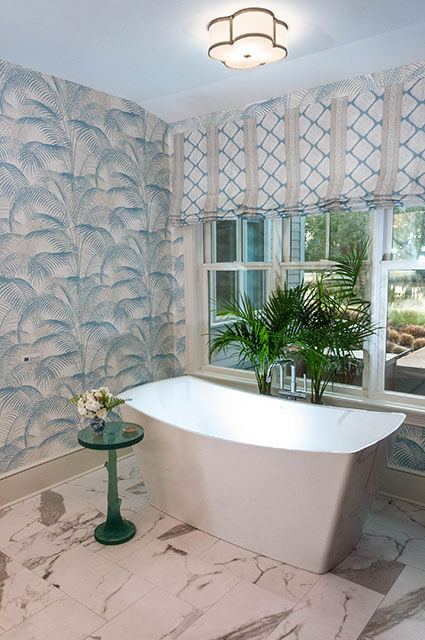 A spacious bathroom featuring a modern white freestanding soaking tub. The walls are covered in light blue palm leaf wallpaper, and a window with a geometric-patterned Roman shade overlooks a garden. A small green accent table holds a vase of flowers next to the tub.