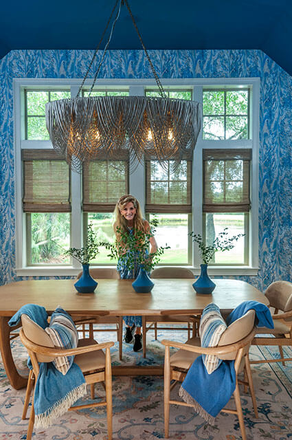 A dining room featuring a long light-wood table and mid-century modern chairs. A large, dramatic beaded chandelier hangs above the table, which is decorated with three vibrant blue vases. The walls are covered in a blue-and-white botanical wallpaper.