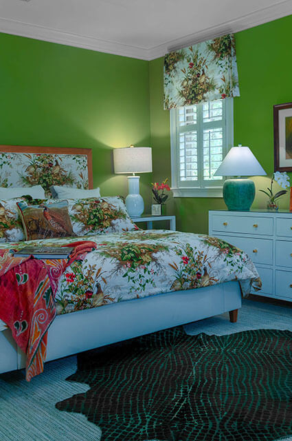 A vibrant green bedroom with a bird-and-botanical print duvet and matching headboard. A turquoise lamp sits on a white dresser next to a window with white shutters. A dark, patterned animal-hide rug is centered on the light blue floor.