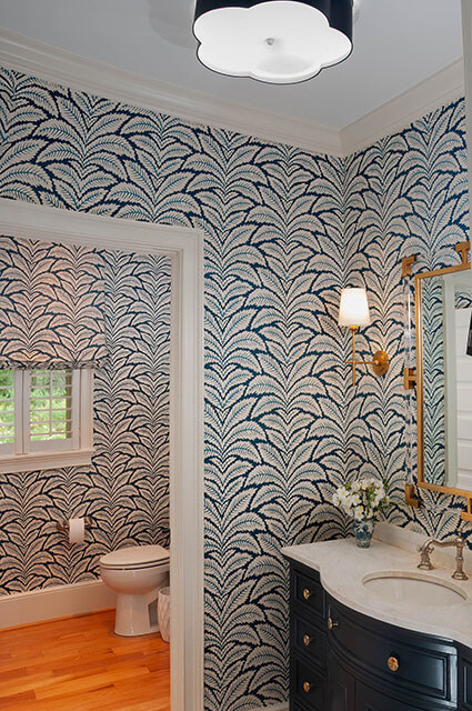 A bathroom featuring bold, navy-and-white palm frond wallpaper. It includes a dark wood vanity with a marble top and a gold-framed mirror with clear acrylic accents. A flower-shaped flush-mount light fixture illuminates the space.