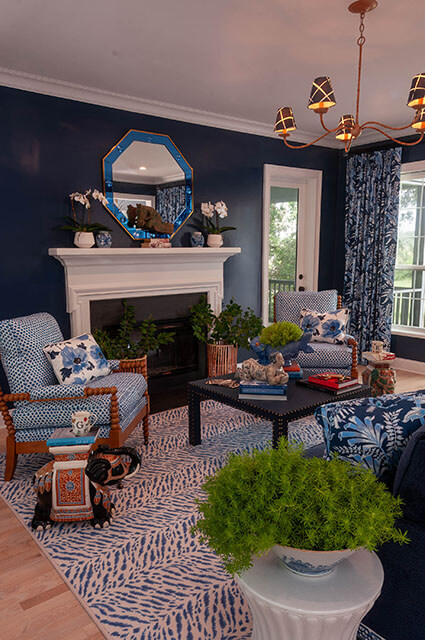 A sophisticated living room with deep navy blue walls and a white fireplace mantle. An octagonal blue-framed mirror hangs above the mantle. The space is filled with patterns, including a blue-and-white zebra-style rug, polka-dot armchairs, and floral pillows.