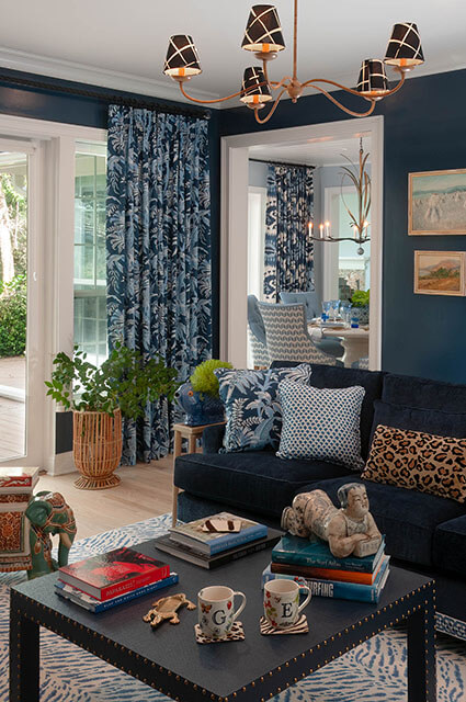 A view of a navy blue living room showing a dark velvet sofa with leopard print and polka-dot pillows. A black studded coffee table holds stacks of books and decorative objects. A modern chandelier with black-and-white patterned shades hangs from the ceiling.