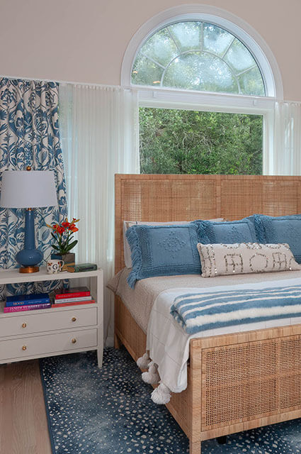 A sunlit bedroom featuring a woven rattan bed frame topped with blue textured pillows and a striped throw. A large arched window looks out onto lush green trees. A blue gourd lamp and vibrant orange flowers sit on a white nightstand next to the bed.