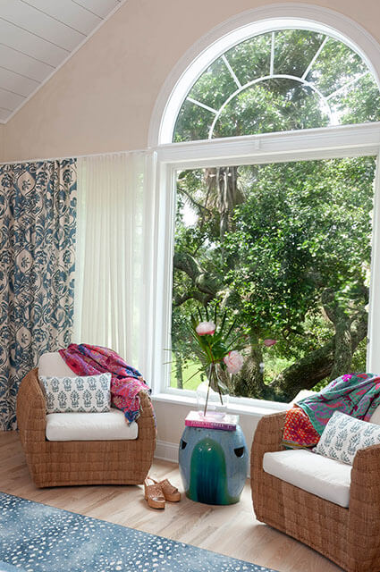A bright seating nook with two large woven armchairs positioned in front of a wide arched window. Colorful paisley and block-print throws are draped over the chairs. A tall blue-and-green glazed ceramic garden stool serves as a side table between them.