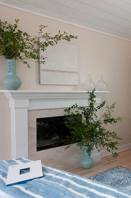 A bright living room featuring a white traditional fireplace mantel with a light stone surround. Above the mantel, a neutral square abstract painting is centered between two clear glass vases. Two large, soft-blue ceramic vases filled with tall green branches flank the fireplace, and a blue-and-white striped upholstered ottoman sits in the foreground.