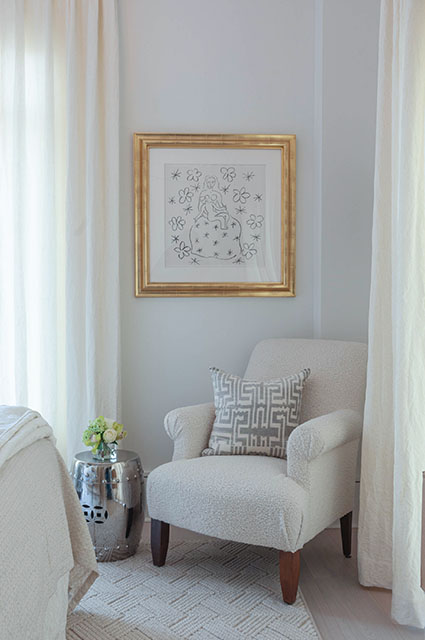 An elegant seating area featuring a cream-colored sherpa or bouclé armchair accented with a grey-and-white geometric Greek key pillow. A gold-framed sketch of a seated figure hangs on the light grey wall above the chair. The space is framed by floor-to-ceiling white curtains, a textured neutral rug, and a polished silver garden stool used as a side table.