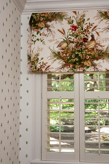 A bright interior corner showcasing a window with white wooden shutters and a decorative fabric valance featuring a classic bird and floral print in red, green, and brown tones. The adjacent wall is covered in a delicate white wallpaper with a small, recurring green leaf pattern, topped with traditional white crown molding.