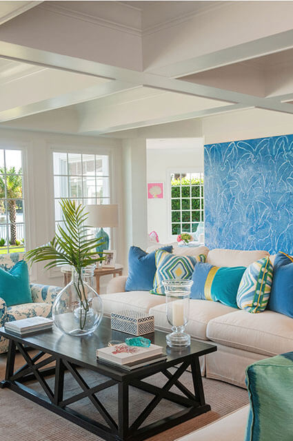 A bright, airy living space with a white sectional sofa featuring a mix of turquoise, yellow, and blue patterned pillows. A large blue-and-white botanical-print wall panel serves as a backdrop. In the foreground, a dark wood X-base coffee table holds a large glass vase with a palm frond and several decorative books, all under a white coffered ceiling.