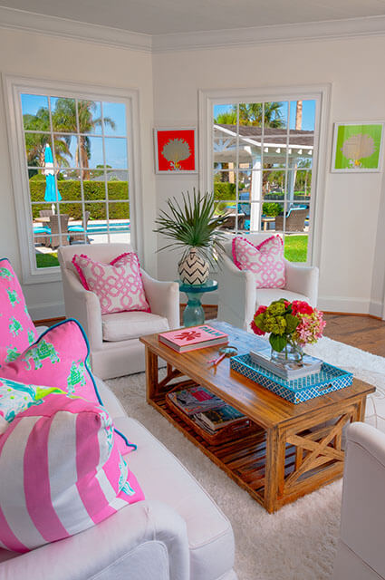 A sunlit room featuring two white armchairs with pink-and-white trellis-patterned pillows. A rustic wooden coffee table sits on a plush white shag rug, topped with a bright blue tray, pink books, and fresh flowers. Through large windows, a view of a patio, pool, and palm trees creates a tropical Florida-style backdrop.