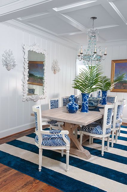 A formal dining room with white vertical shiplap walls and a coffered ceiling. A light wood trestle table is surrounded by white chairs upholstered in a bold blue-and-white zebra print. A large navy-and-white striped rug anchors the space, while a blue glass chandelier and coral-shaped wall sconces add coastal flair.