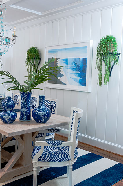 A close-up view of a coastal dining setup featuring blue-and-white cherry blossom patterned porcelain vases on a light wood table. The white shiplap wall behind is decorated with a blue-and-white abstract beach landscape painting and two black wall brackets holding cascading green plants.