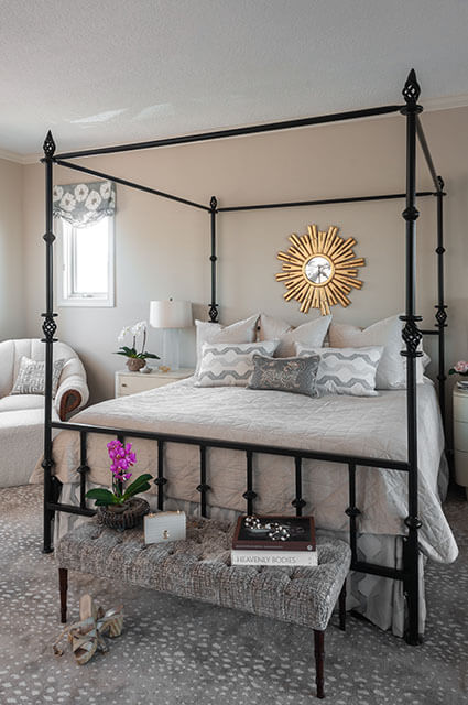 A luxurious master bedroom featuring a black iron four-poster canopy bed with neutral quilted bedding and grey-and-white patterned accent pillows. Above the headboard hangs a large, ornate gold sunburst mirror. In the foreground, a tufted grey upholstered bench sits on a grey antelope-print carpet, accented with a purple orchid and coffee table books.