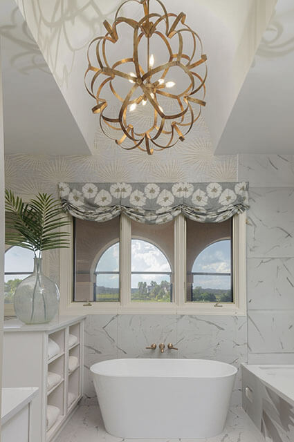 A bright, high-end bathroom featuring a freestanding white soaking tub set against white marble-patterned wall tiles. Above the tub, a window with arched panes is dressed in a grey-and-white floral Roman shade. A dramatic, large gold orbital ribbon chandelier hangs from the ceiling, while a glass vase with a palm frond sits on a nearby white vanity.