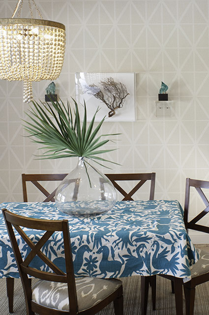 Dining area with a blue and white otomi-print tablecloth, cross-back wood chairs, large glass demijohn vase with palm frond, beaded wood chandelier, and geometric wallpaper with framed coral and branch wall art