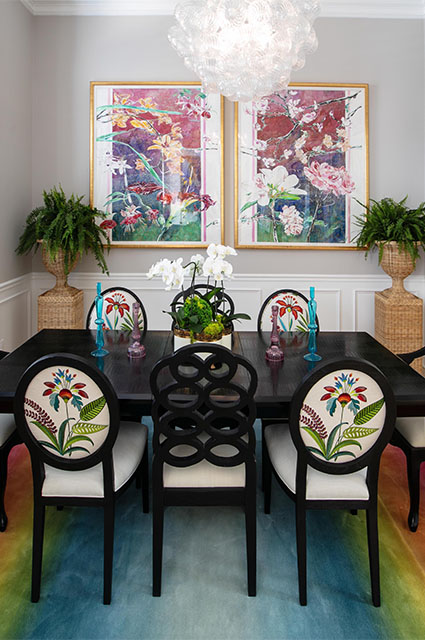 A sophisticated dining room featuring a dark wood table and black oval-back chairs with custom botanical embroidery. Two large, gold-framed floral artworks dominate the grey wall. A unique glass bubble chandelier hangs above the table, which is set with colorful glassware and orchid centerpieces, flanked by a floral-patterned window valance.